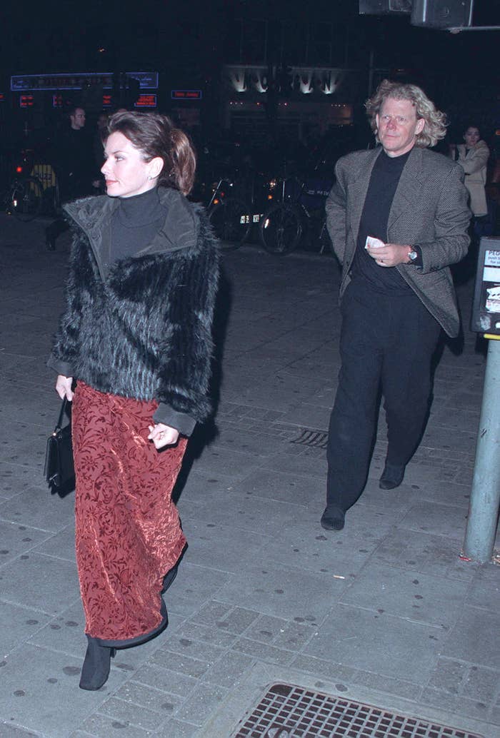 Shania Twain is walking on a city street, wearing a fur jacket and patterned skirt, followed by Mutt Lange