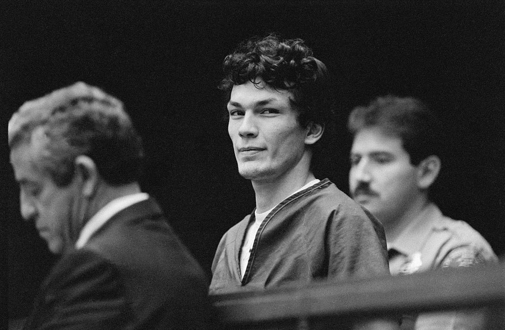 Three men are in a courtroom. One, Richard Ramirez, in a prison jumpsuit, looks toward the camera, while the other two men stand beside and behind him