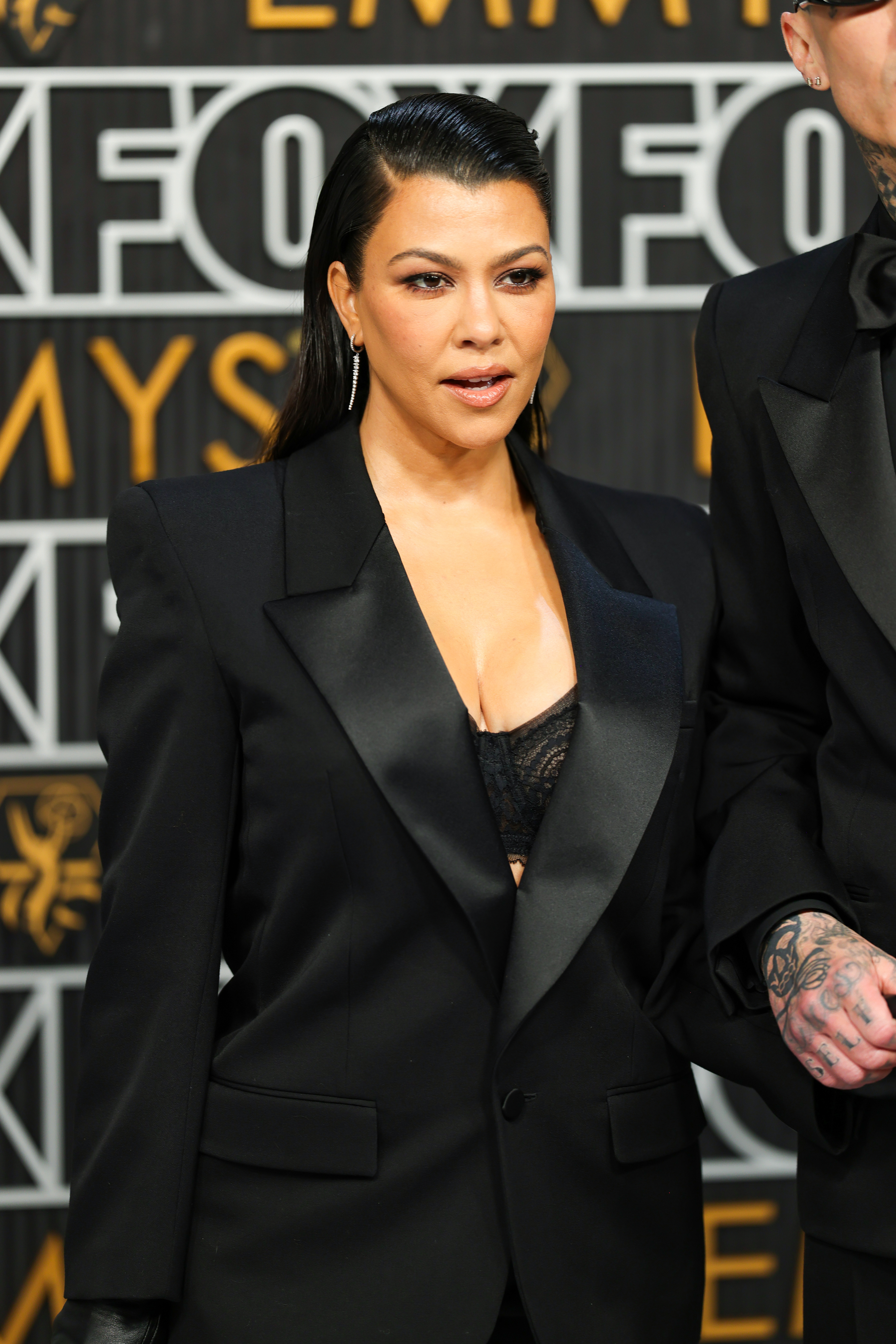 Kourtney Kardashian wearing a tailored blazer with satin lapels and a bra top, standing next to her husband Travis