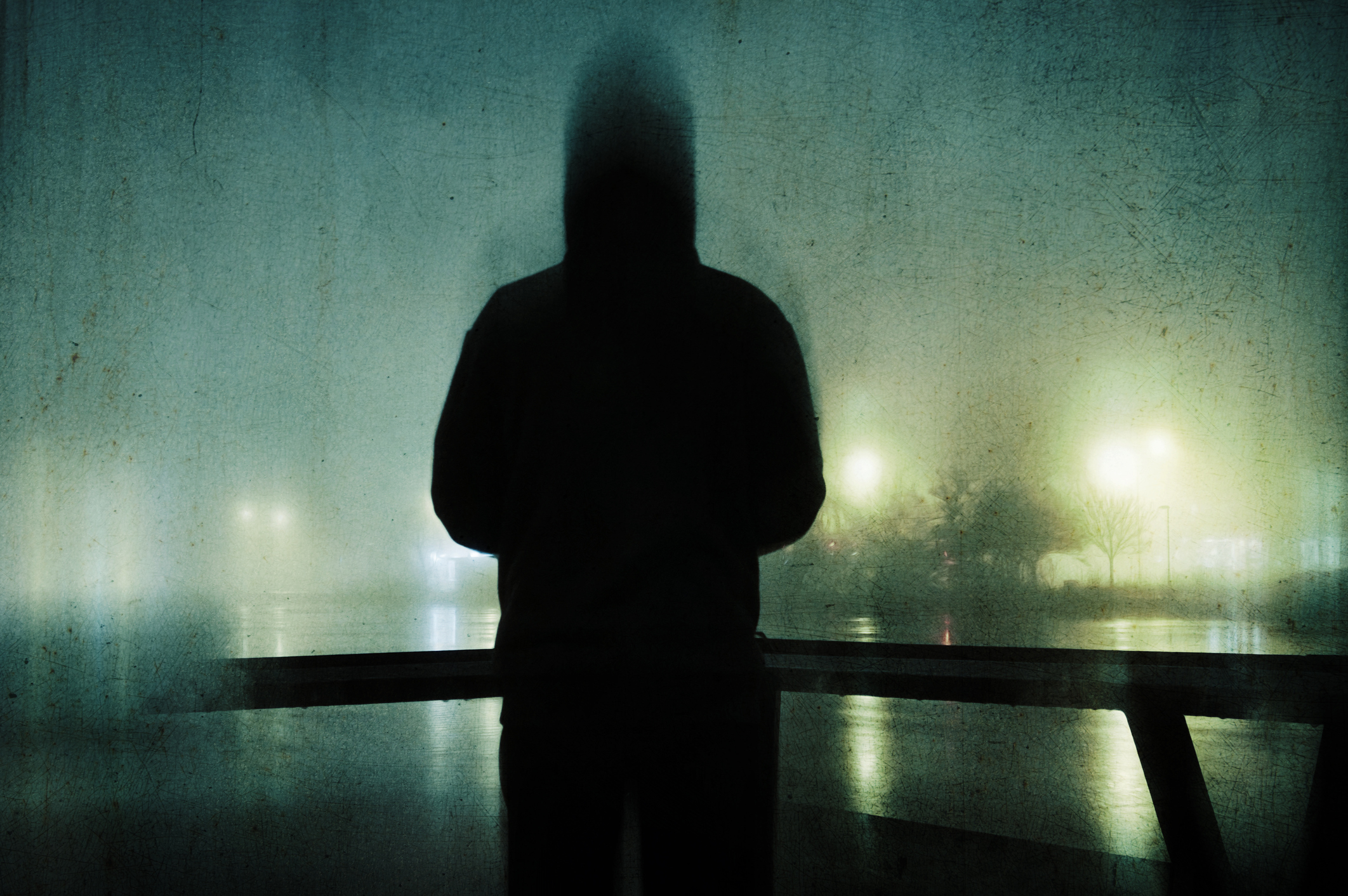 The image shows the silhouette of a person wearing a hooded jacket, standing in a foggy, dimly-lit street at night