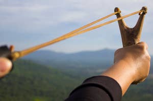 A hand holding a slingshot aims at an unseen target with a mountainous landscape in the background