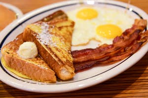 A breakfast plate with French toast topped with butter and powdered sugar, two sunny side up eggs, and three strips of bacon