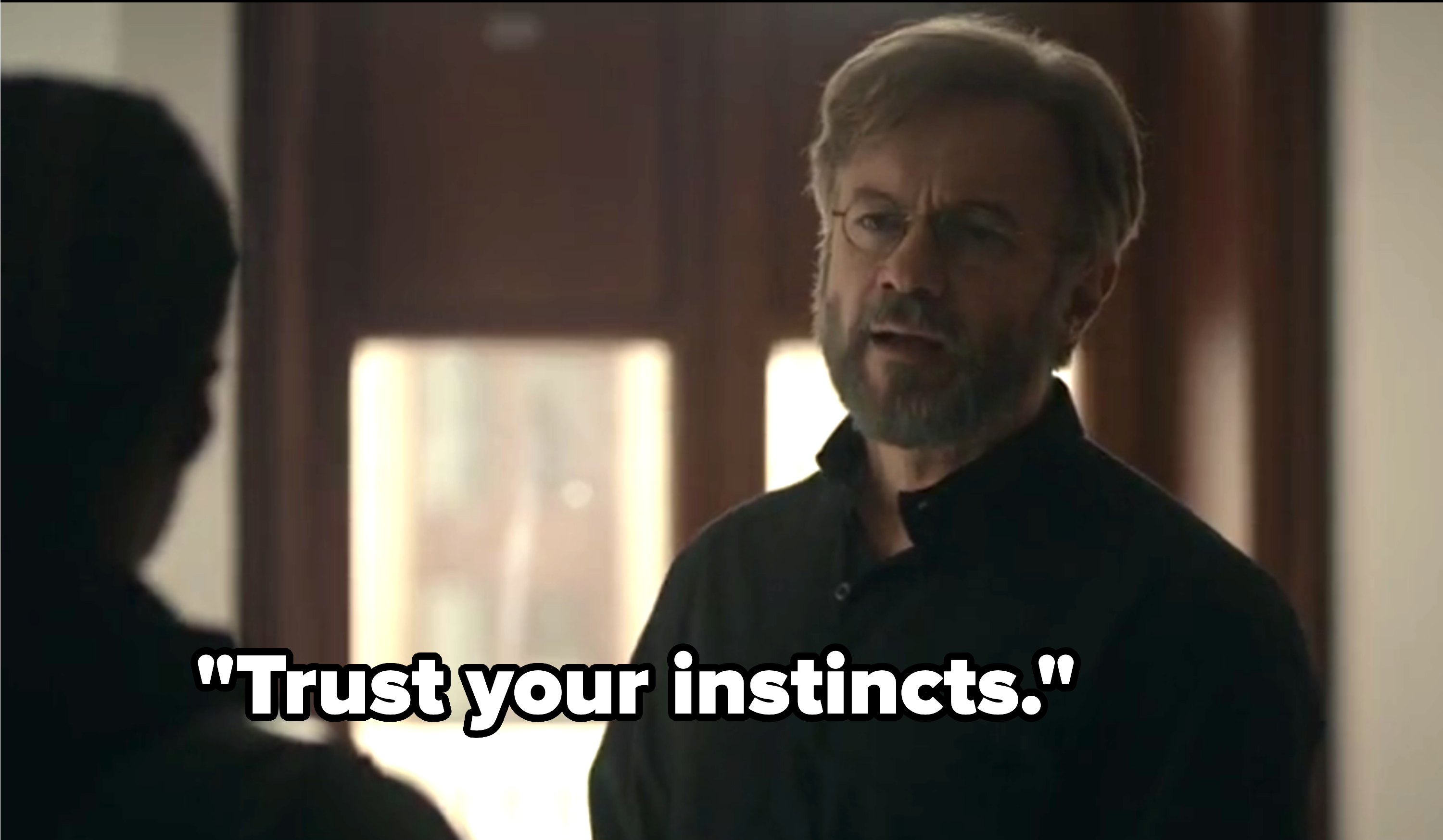 A man with glasses, a beard, and short hair wears a dark shirt and stands facing another person indoors. Their conversation appears serious