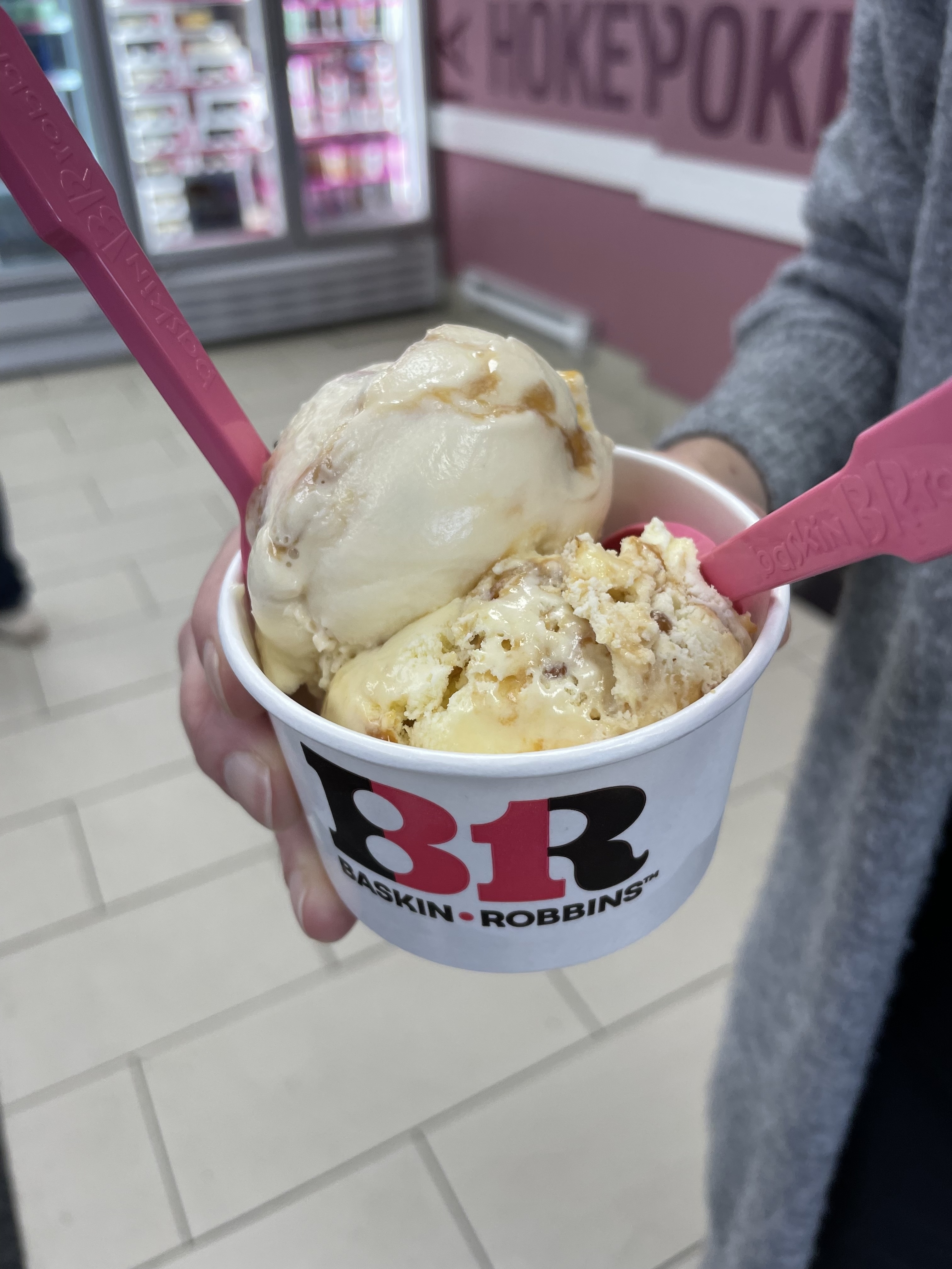 Hand holding a Baskin-Robbins cup with two scoops of ice cream, each topped with sauce. The background shows an ice cream shop interior