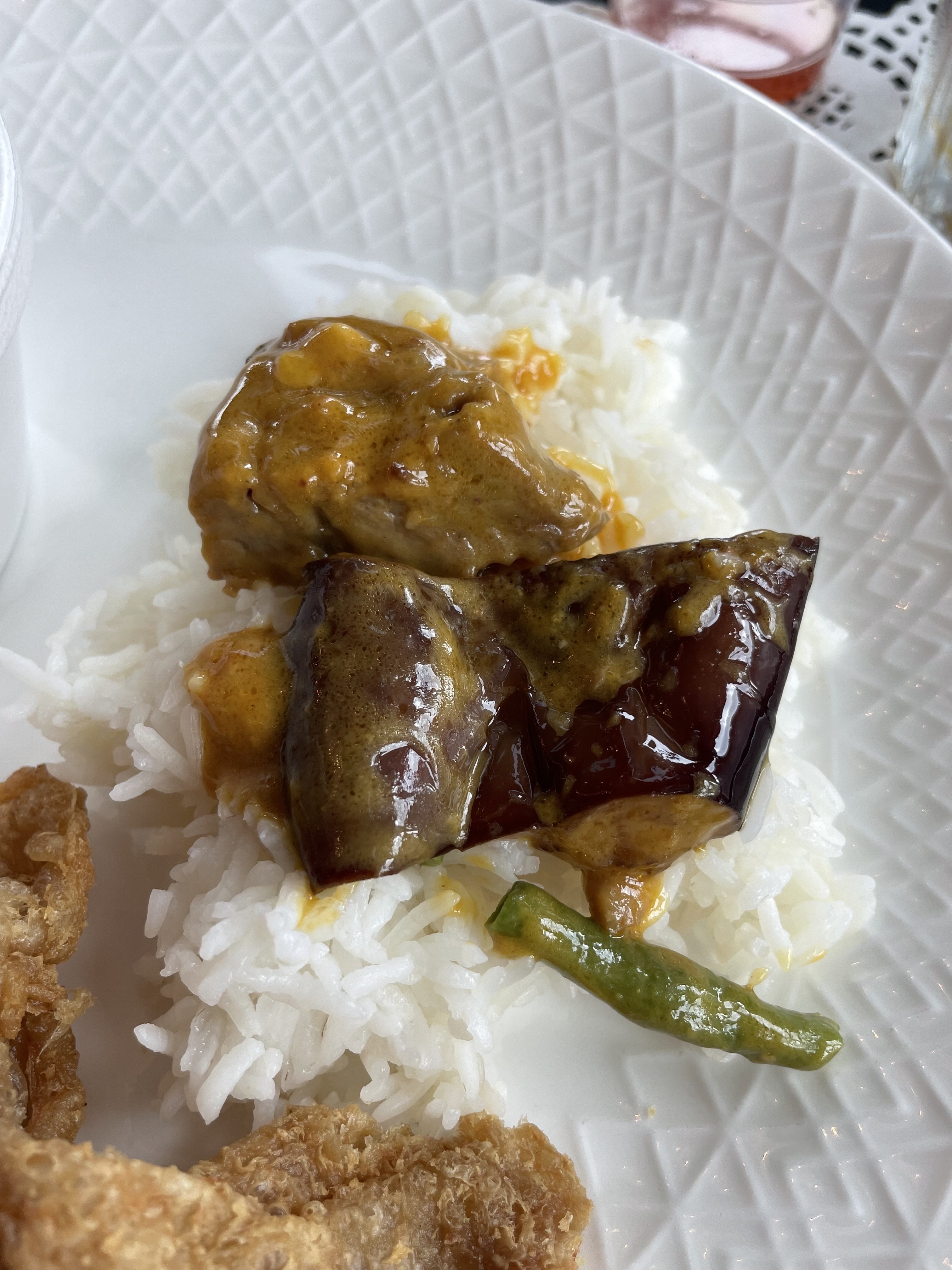 A plate of white rice topped with eggplant and a piece of meat, accompanied by a green pepper and a portion of fried food