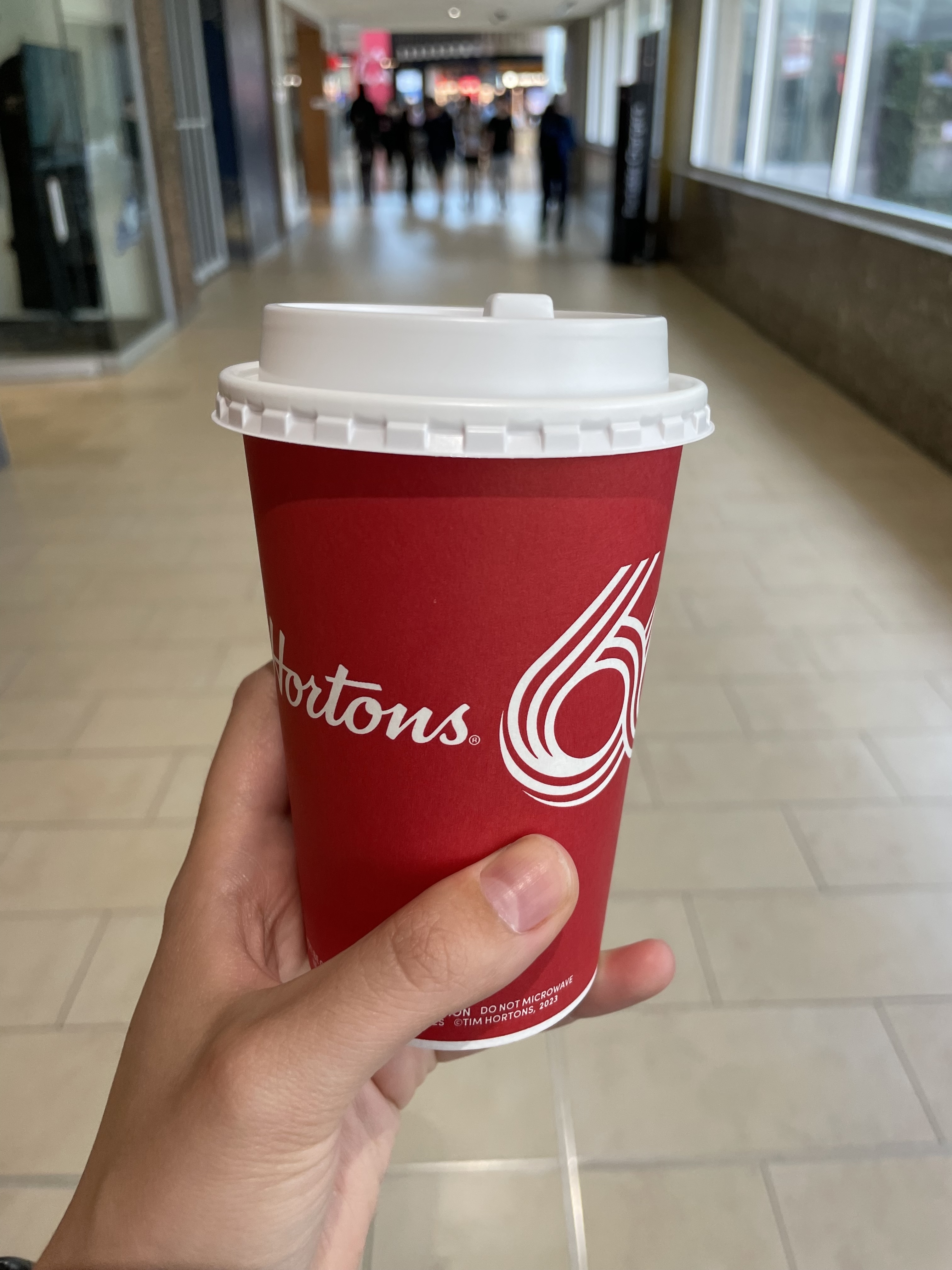 A hand holding a Tim Hortons coffee cup in what appears to be a shopping mall