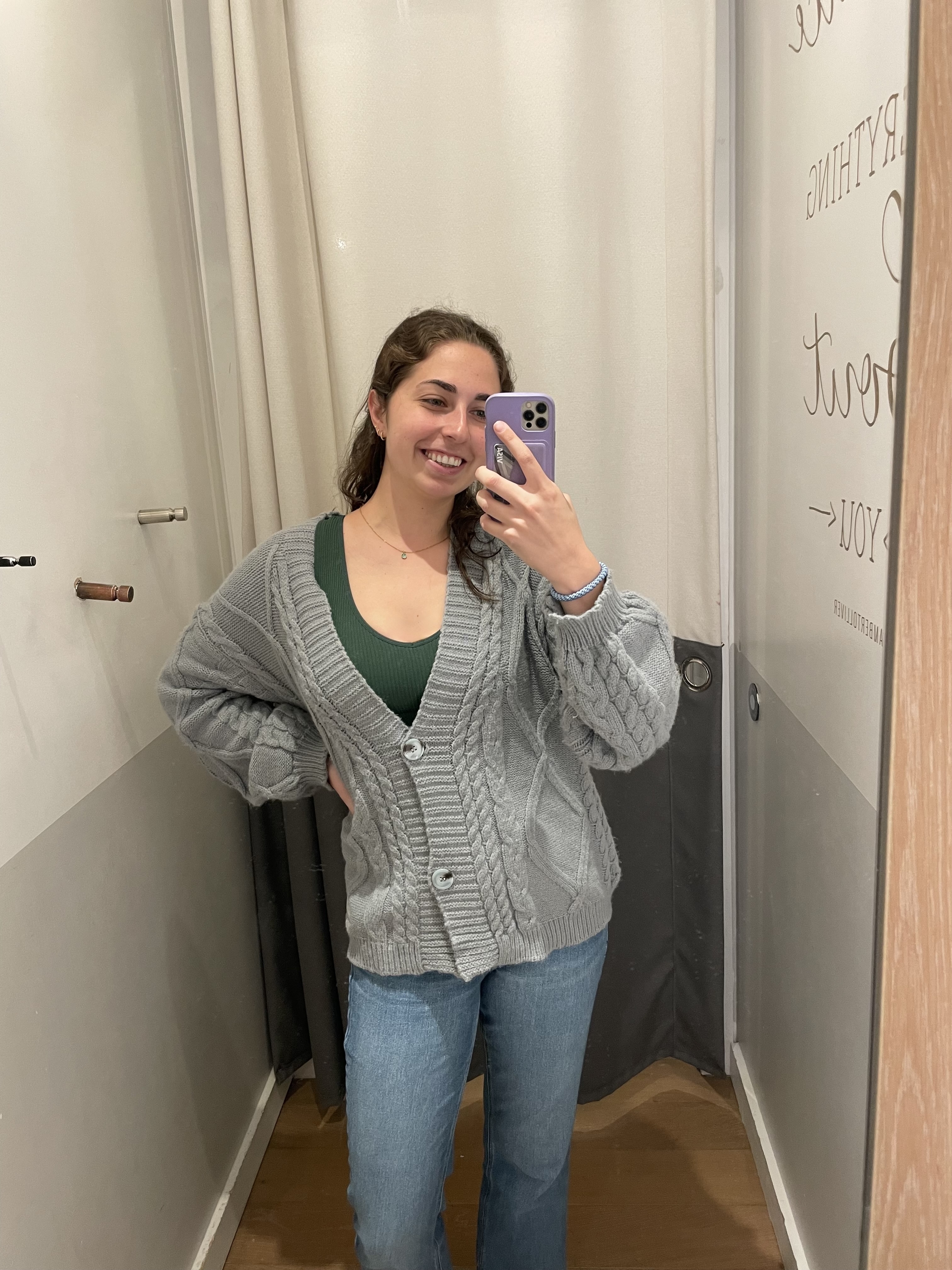 Woman in a cozy gray cardigan over a green top, smiling and taking a mirror selfie in a fitting room