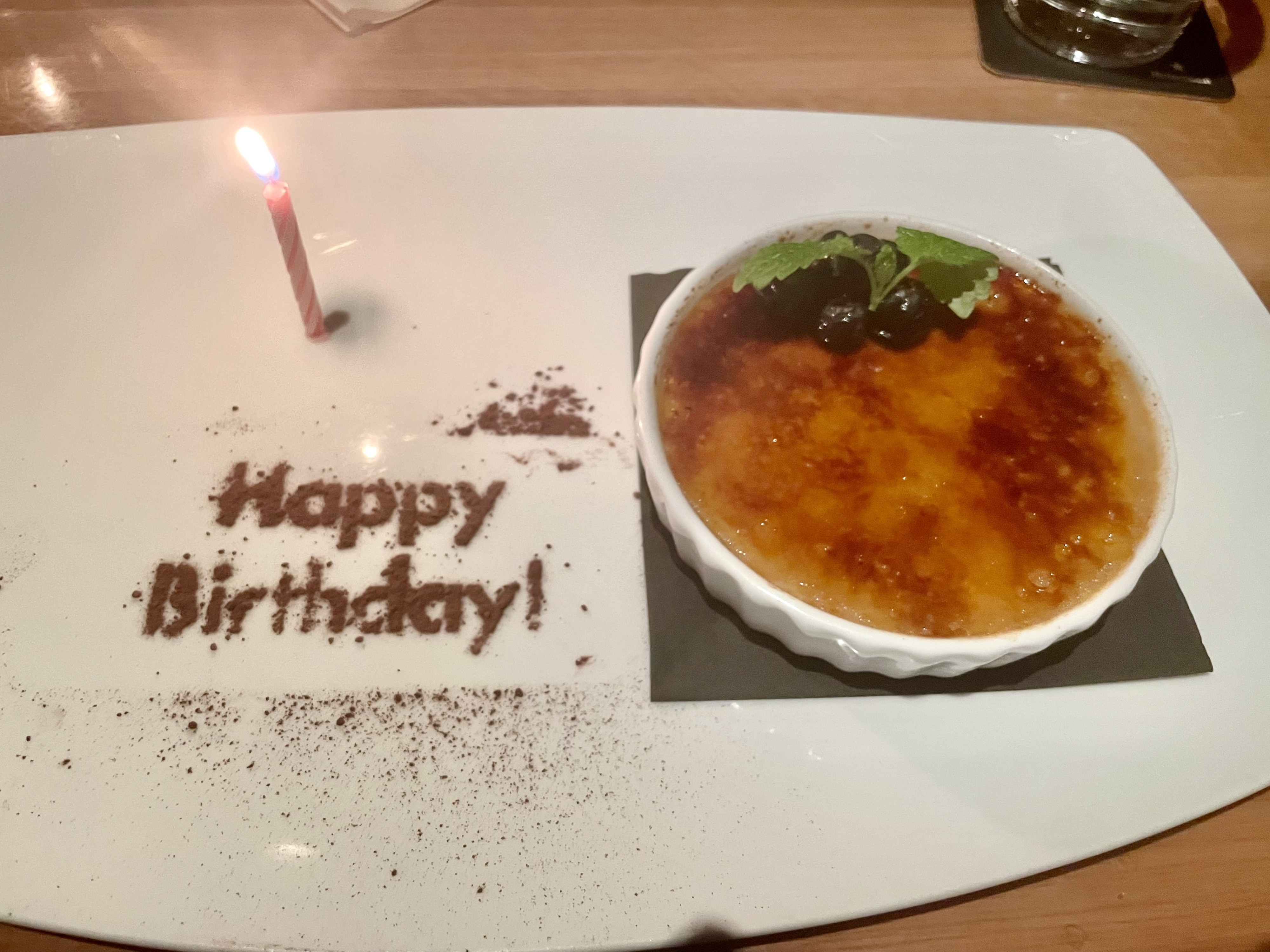 A crème brûlée with a single lit birthday candle, "Happy Birthday!" written in chocolate on the plate
