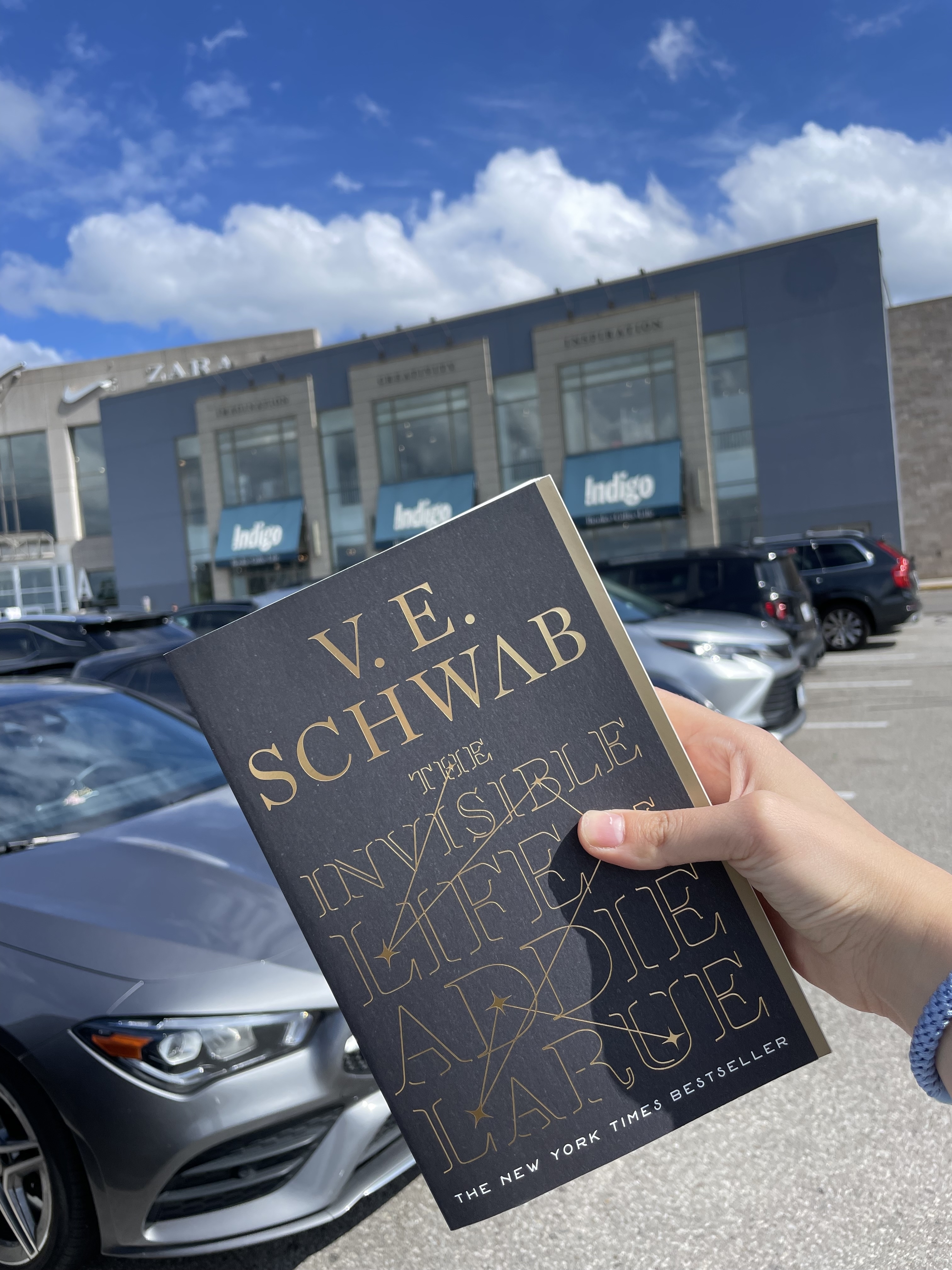 A hand holds a copy of "The Invisible Life of Addie LaRue" by V. E. Schwab in a parking lot with a Zara and Indigo store in the background