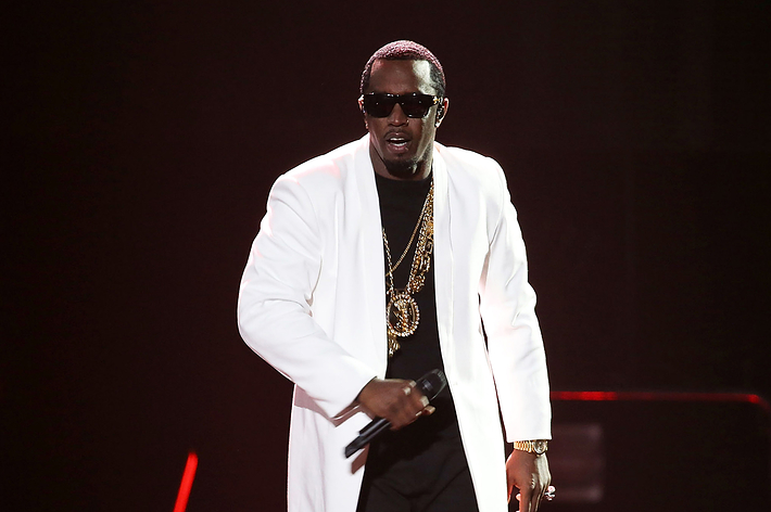Sean "Diddy" Combs performing on stage wearing a white coat over a black outfit, accessorized with sunglasses and gold chains, holding a microphone