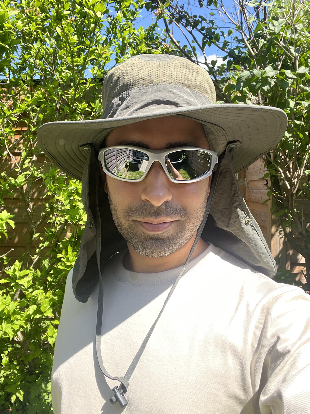 Person in a wide-brim hat with neck flap, reflective sunglasses, and a light shirt stands outdoors with green foliage in the background. Name unknown