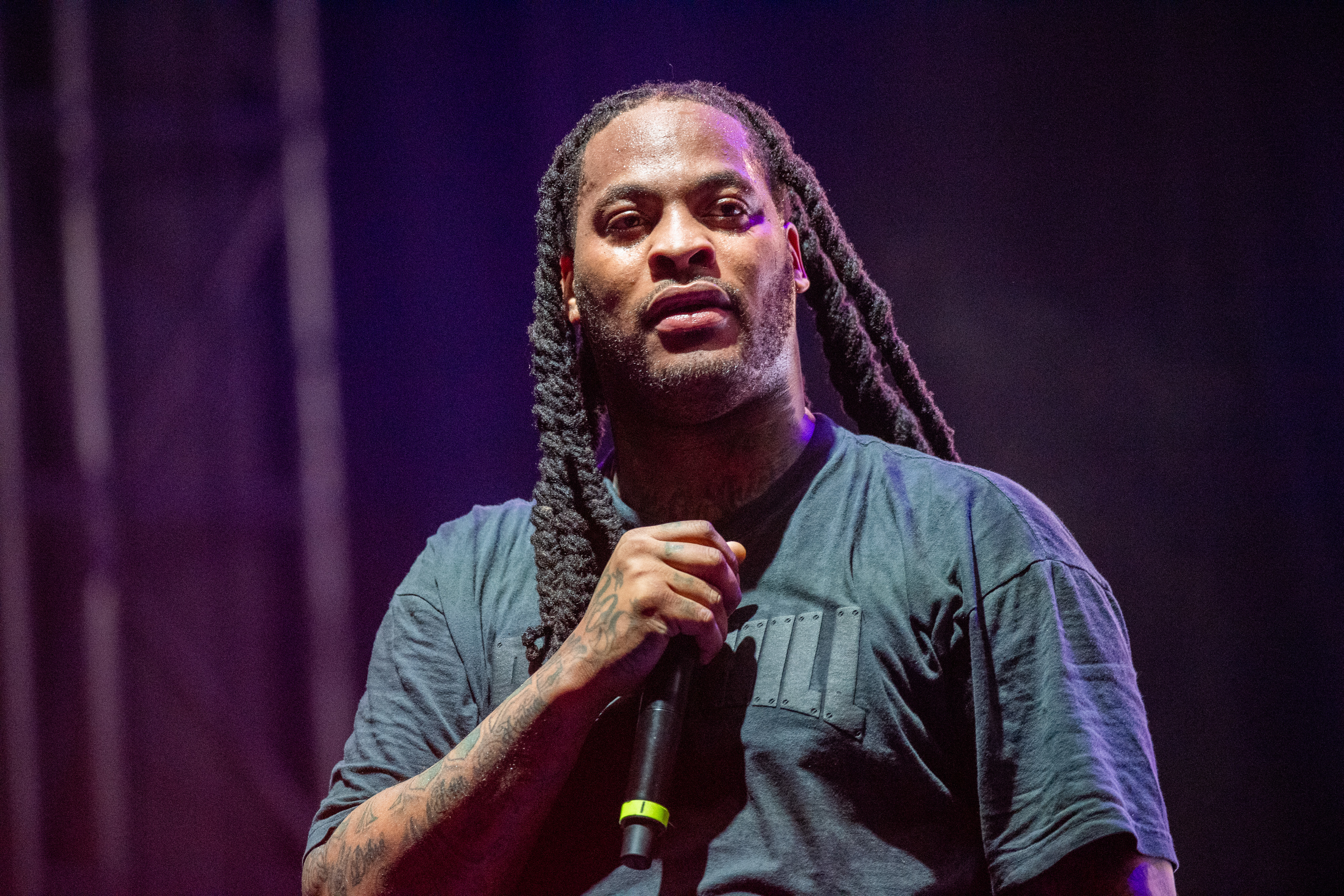 Waka Flocka Flame is holding a microphone on stage, wearing a casual t-shirt, with long dreadlocks over his shoulders