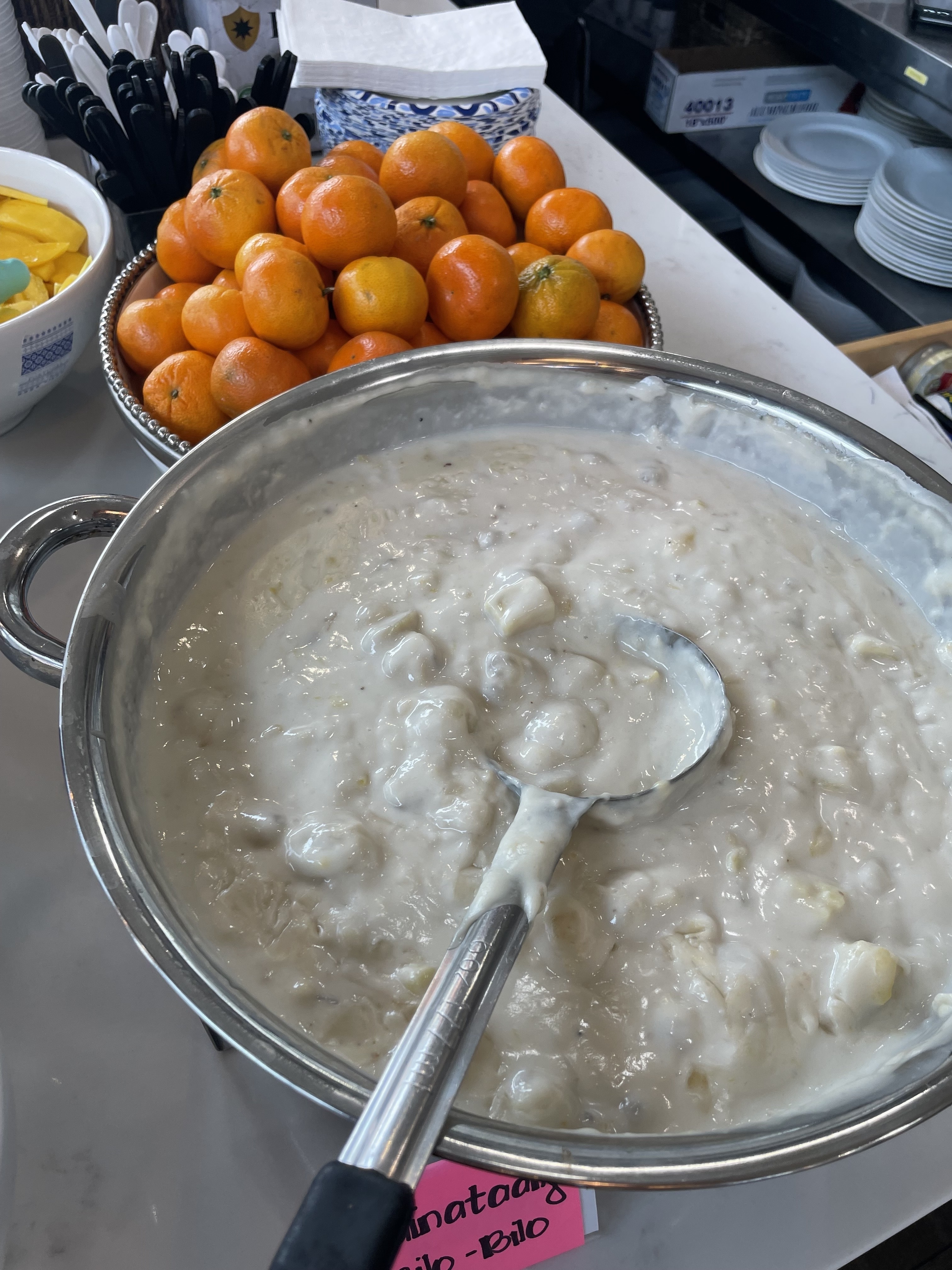 A large bowl of rice pudding with a serving spoon next to a bowl of fresh oranges. A pink label reads "Banana Pudding."