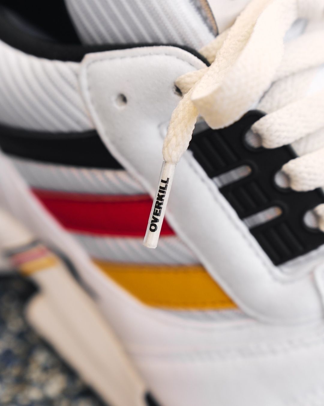 Close-up of a sneaker with a white lace featuring the text &quot;OVERKILL&quot; on the lace tip. The sneaker has black, red, and yellow accents