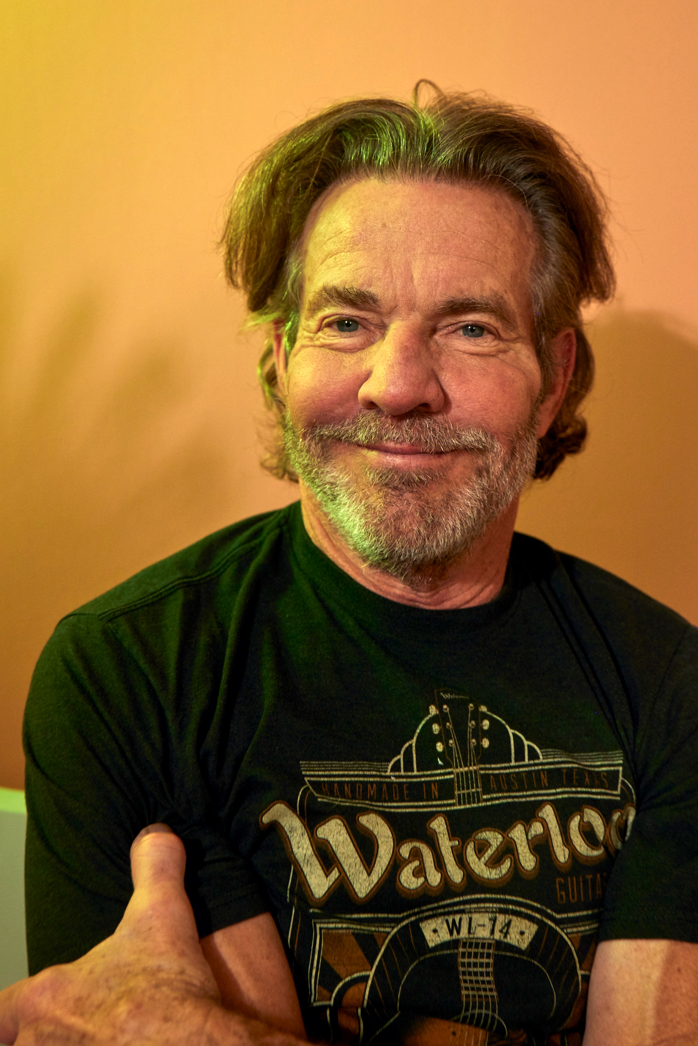 Dennis Quaid wearing a casual &quot;Waterloo Guitars&quot; t-shirt, smiling and posing indoors