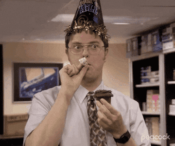 Dwight Schrute wears a party hat, blows a party horn, and holds a piece of chocolate cake in "The Office."