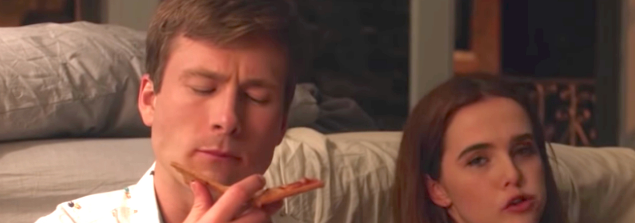 Glen Powell and Zoey Deutch sit closely, each holding a slice of pizza, with Glen looking contemplative and Zoey talking as Charlie and Harper in Set It Up