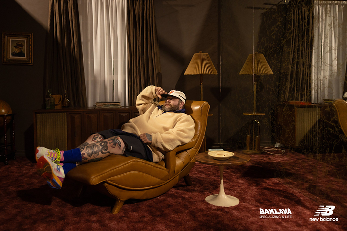 Man lounging in a vintage-style room, wearing New Balance sneakers. Room has retro decor, with a brown chair, globe, curtains, lamps, and framed art
