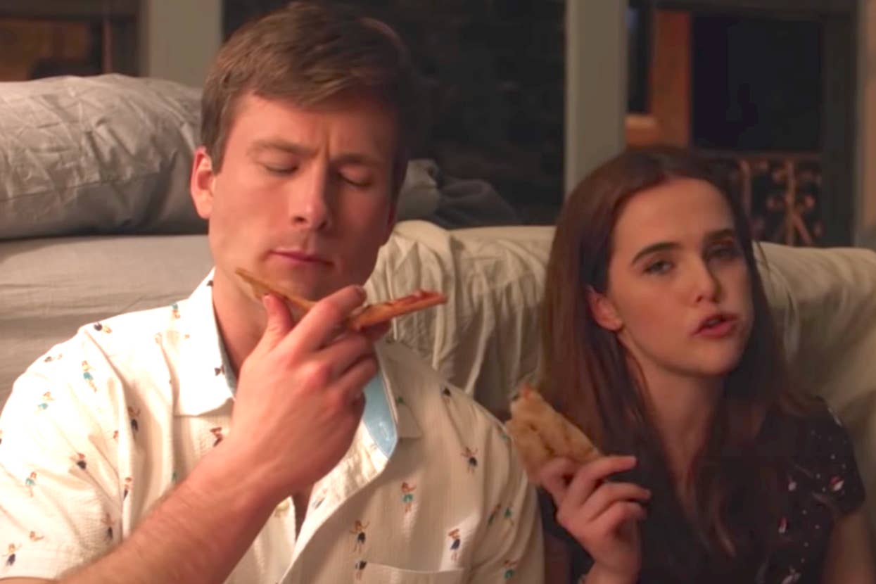 Glen Powell and Zoey Deutch sit closely, each holding a slice of pizza, with Glen looking contemplative and Zoey talking as Charlie and Harper in Set It Up