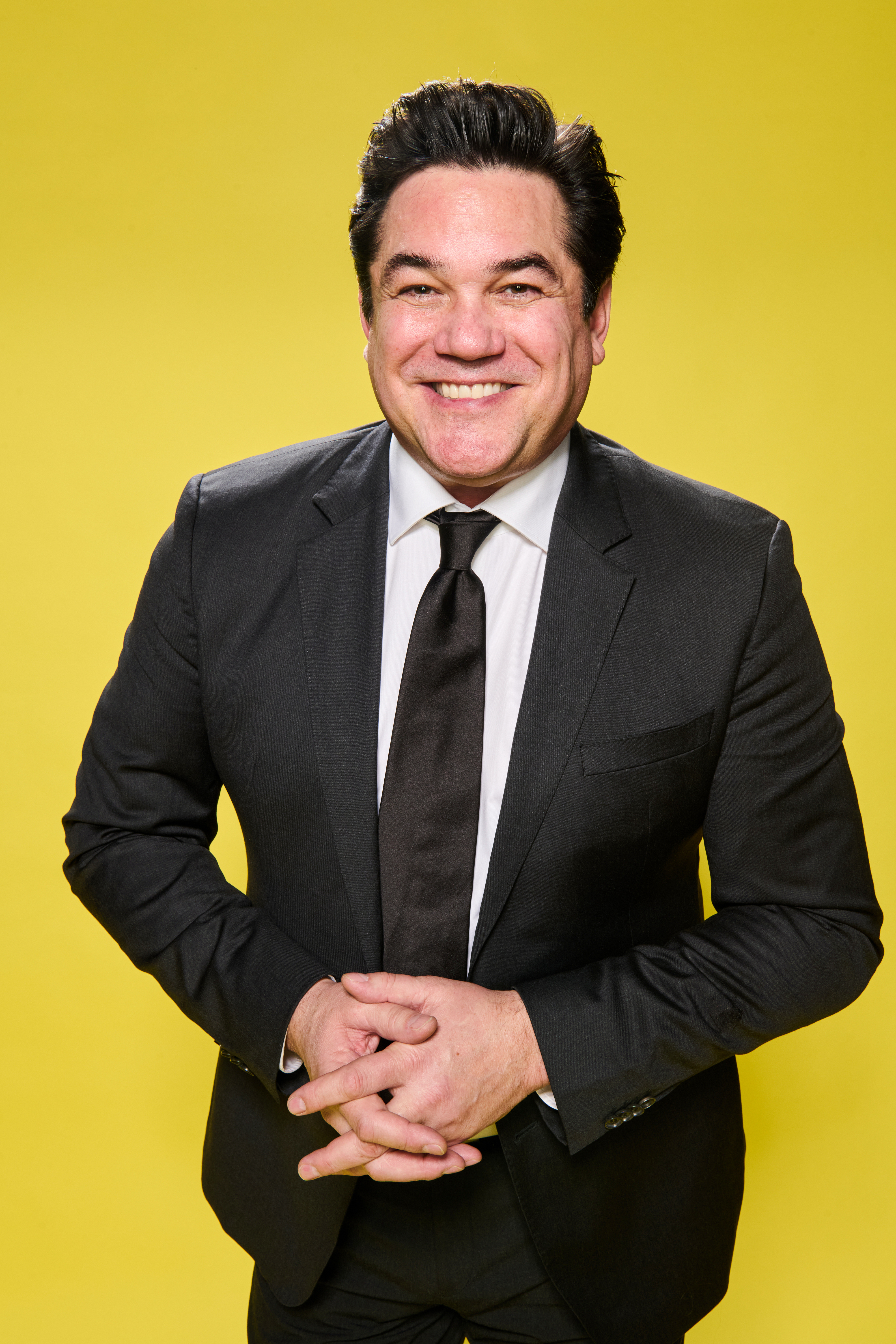 Dean Cain, smiling, poses in a black suit and dark tie against a yellow backdrop