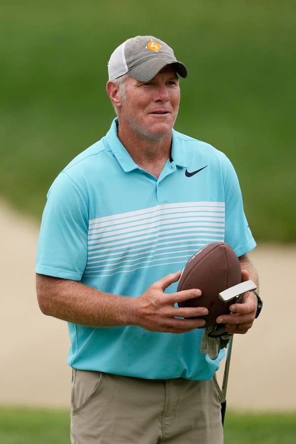 Brett Favre stands outdoors, wearing a polo shirt, cap, and holding a football