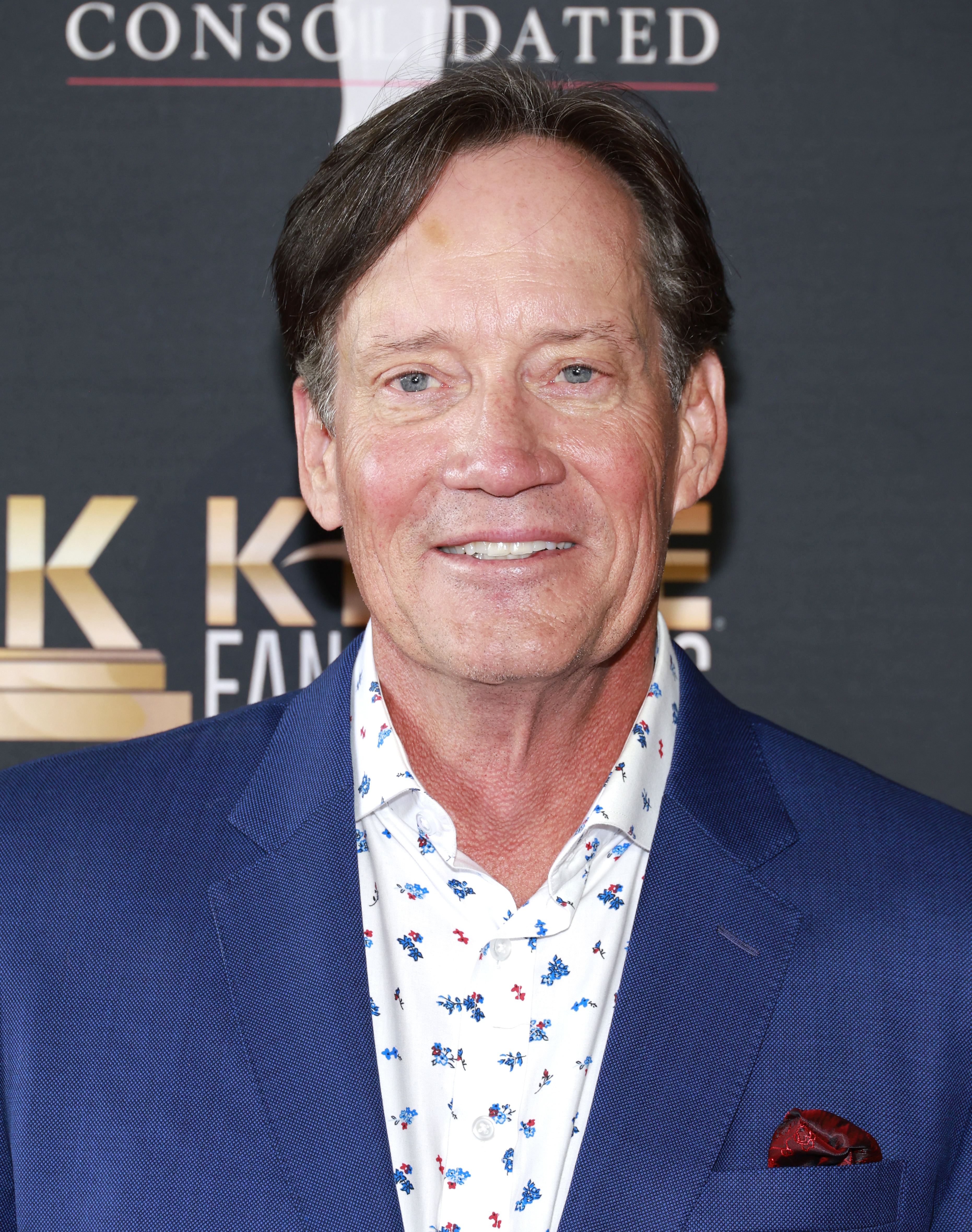 Kevin Sorbo smiles while posing on a red carpet, wearing a blue suit and a white shirt with a colorful pattern