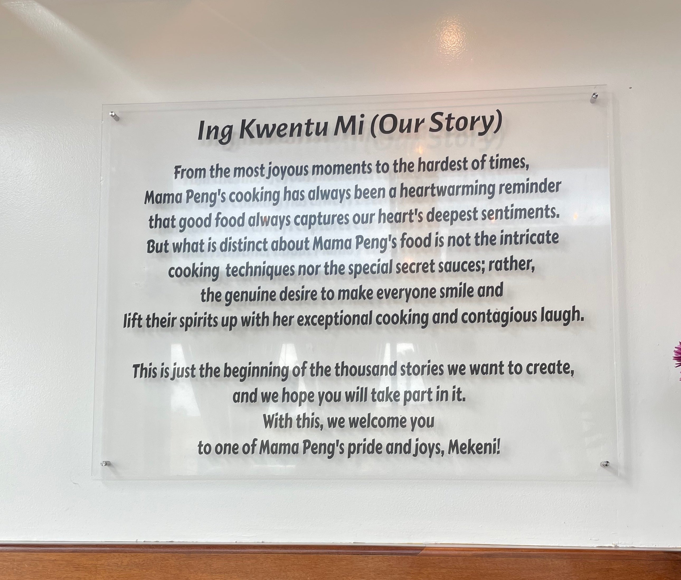 Restaurant wall with a sign summarizing Mama Peng's cooking philosophy and mission, titled "Ing Kwentu Mi (Our Story)." Empty chairs and a table are visible below