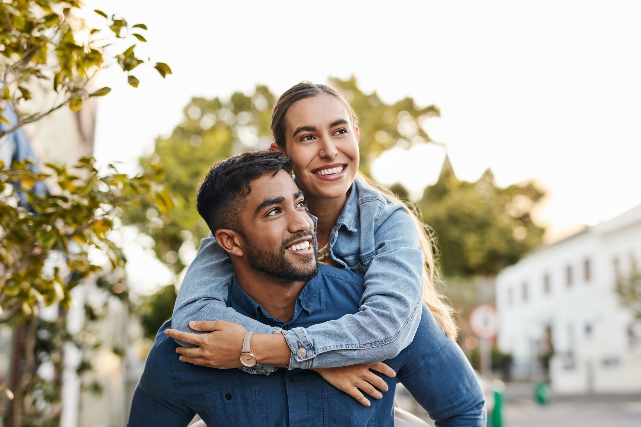 A couple stands outdoors. A man gives a piggyback ride to a smiling woman. Both are wearing casual denim outfits