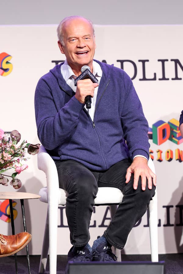 Kelsey Grammer smiles while seated on stage at a podcast event, holding a microphone and wearing casual attire