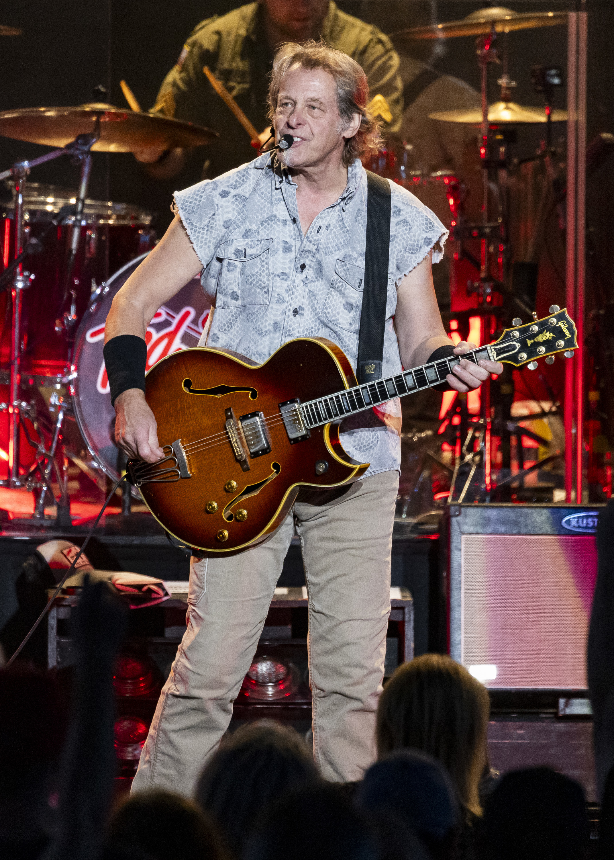 Ted Nugent performs on stage, wearing a sleeveless shirt and beige pants, playing an electric guitar. Drummer is visible in the background