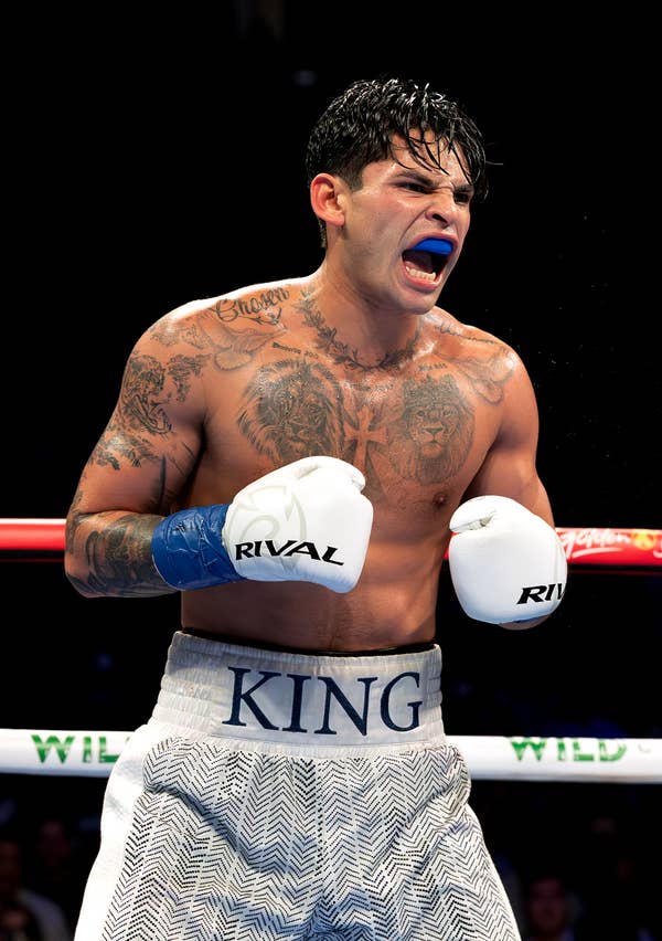 Ryan Garcia in a boxing ring during a match, wearing white trunks with "KING" written on them, mouthguard in place, and tattoos visible on his upper body