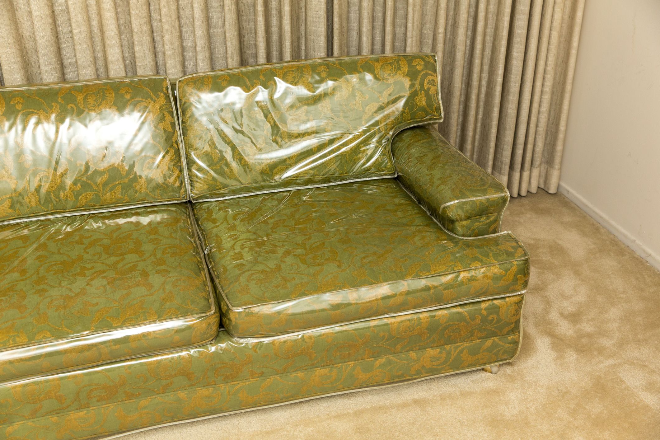 An ornate patterned couch covered with protective plastic is placed on a carpeted floor in front of closed curtains