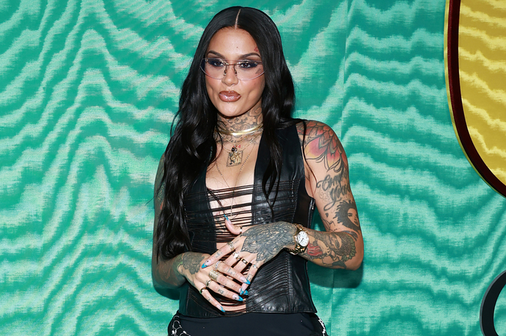 Kehlani in a black strappy top and black pants with visible tattoos on her arms and hands, posing on a patterned backdrop at a music event