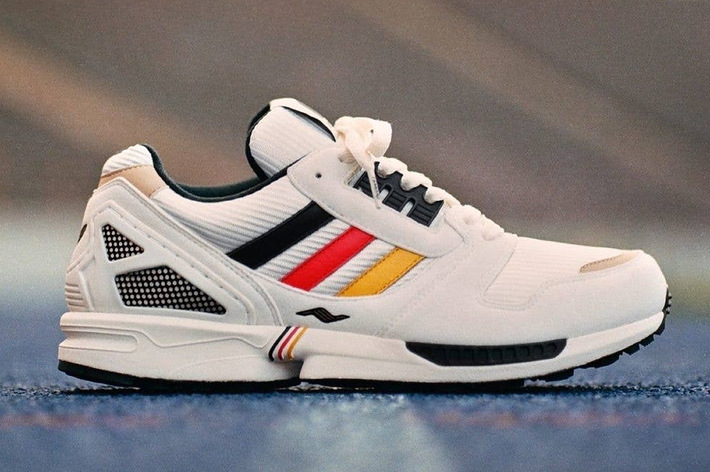 Close-up of an Adidas sneaker with red, yellow, and black stripes on the side, set on an outdoor sports track background