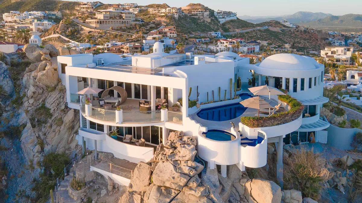 Aerial view of a luxurious cliffside mansion with a stunning pool, terraces, and scenic landscape in a hilly coastal area featured in a travel article