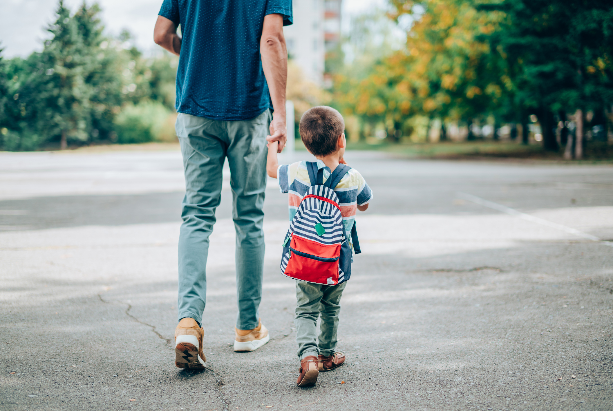 An adult and a child, wearing a backpack, walk hand in hand on an outdoor path surrounded by trees