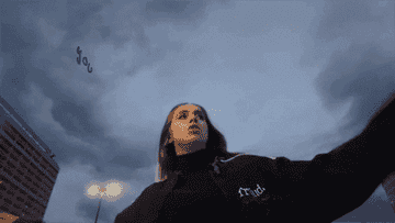 I don't know who this person is. The image shows a person in motion looking up at the sky, wearing a dark jacket that says "Proud." Buildings and streetlights are in the background