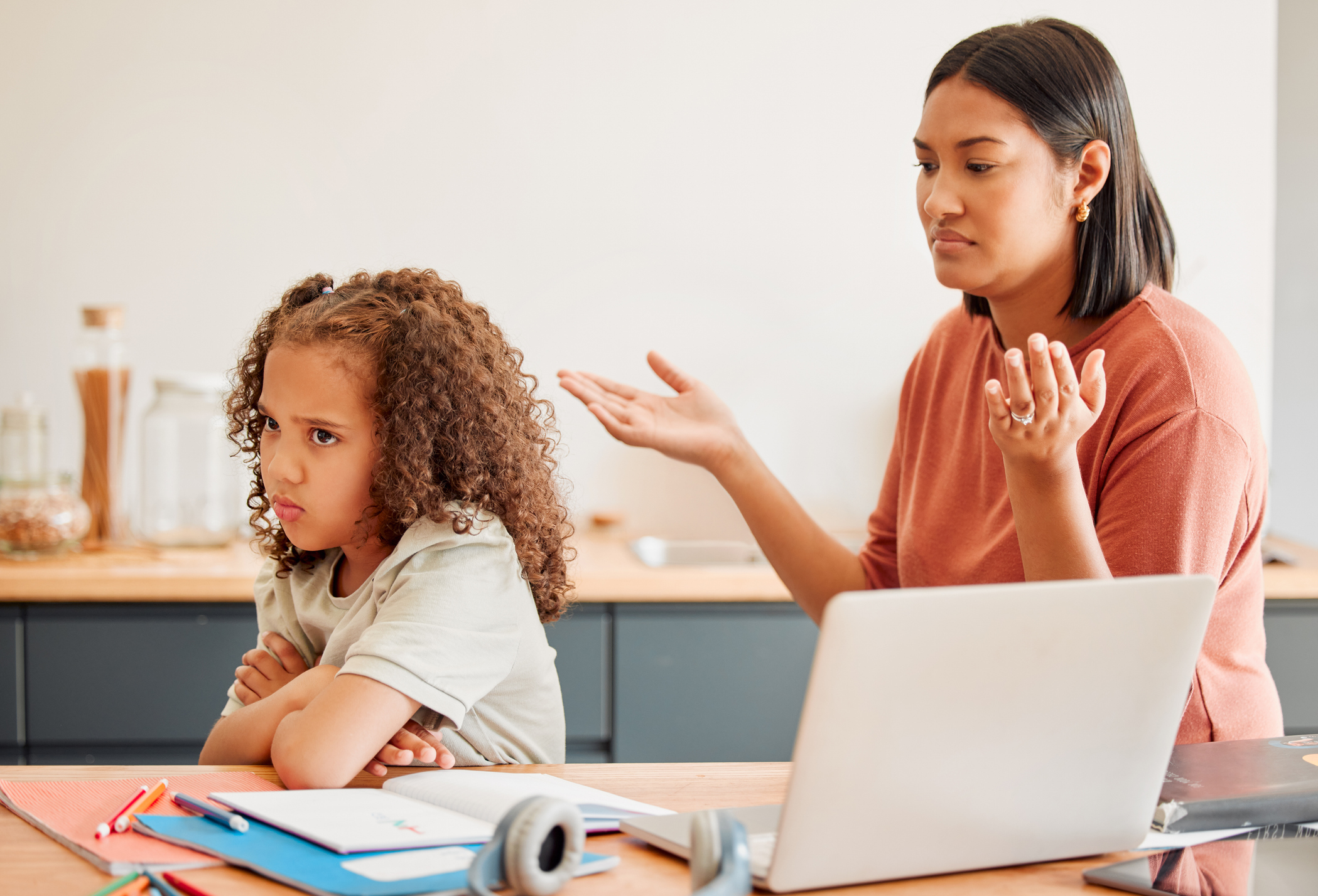 Mother and child engaging in a tense discussion, with the child looking upset and the mother gesturing with frustration at a kitchen table with a laptop and notebooks