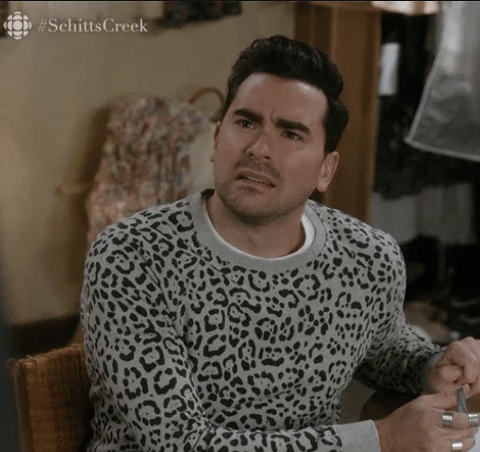 David Rose from Schitt's Creek wears a patterned sweatshirt and sits at a table with a confused expression