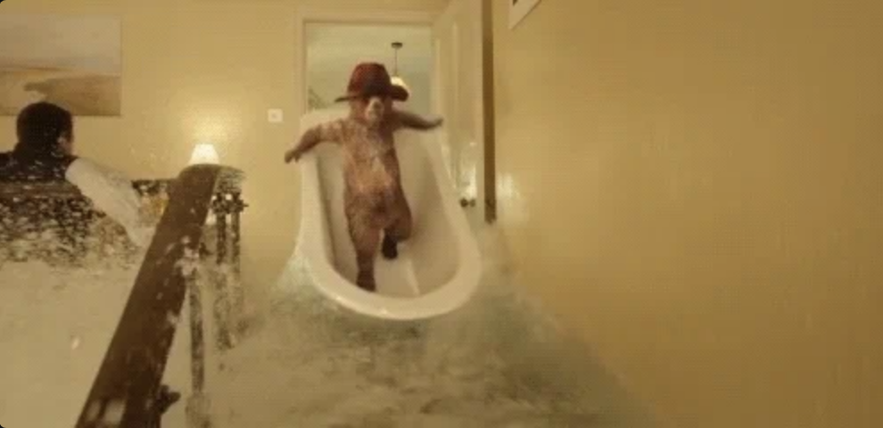 Paddington Bear in a bathtub, wearing a hat, slides down a staircase as water floods the area. Another person is visible to the side, dodging the water