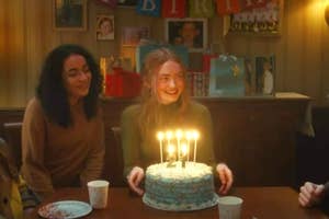 A girl is smiling at her birthday party, holding a cake with lit candles shaped as the number "2". She is surrounded by friends, and there are gifts and decorations behind her