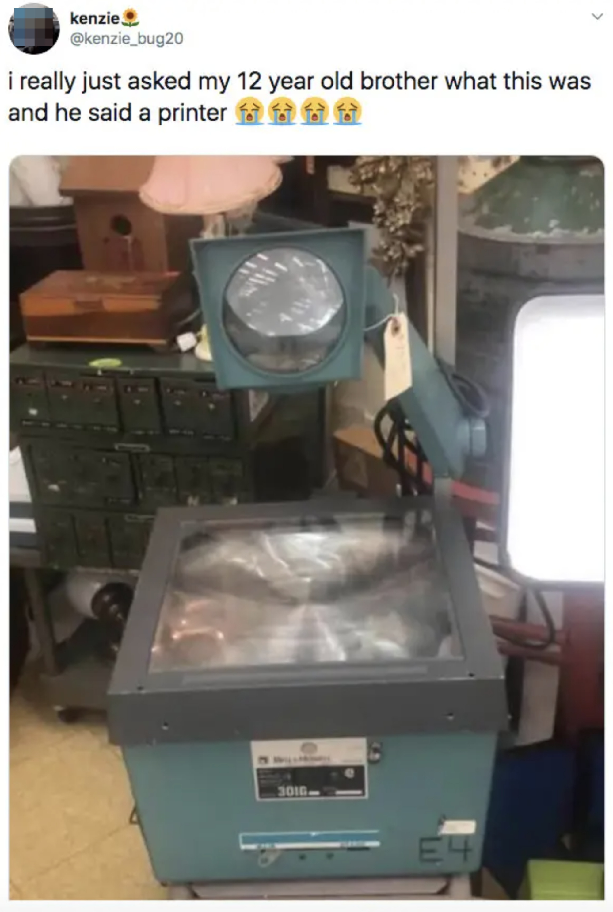 An overhead projector on a desk with a tweet above saying, &quot;i really just asked my 12 year old brother what this was and he said a printer&quot; followed by crying emojis
