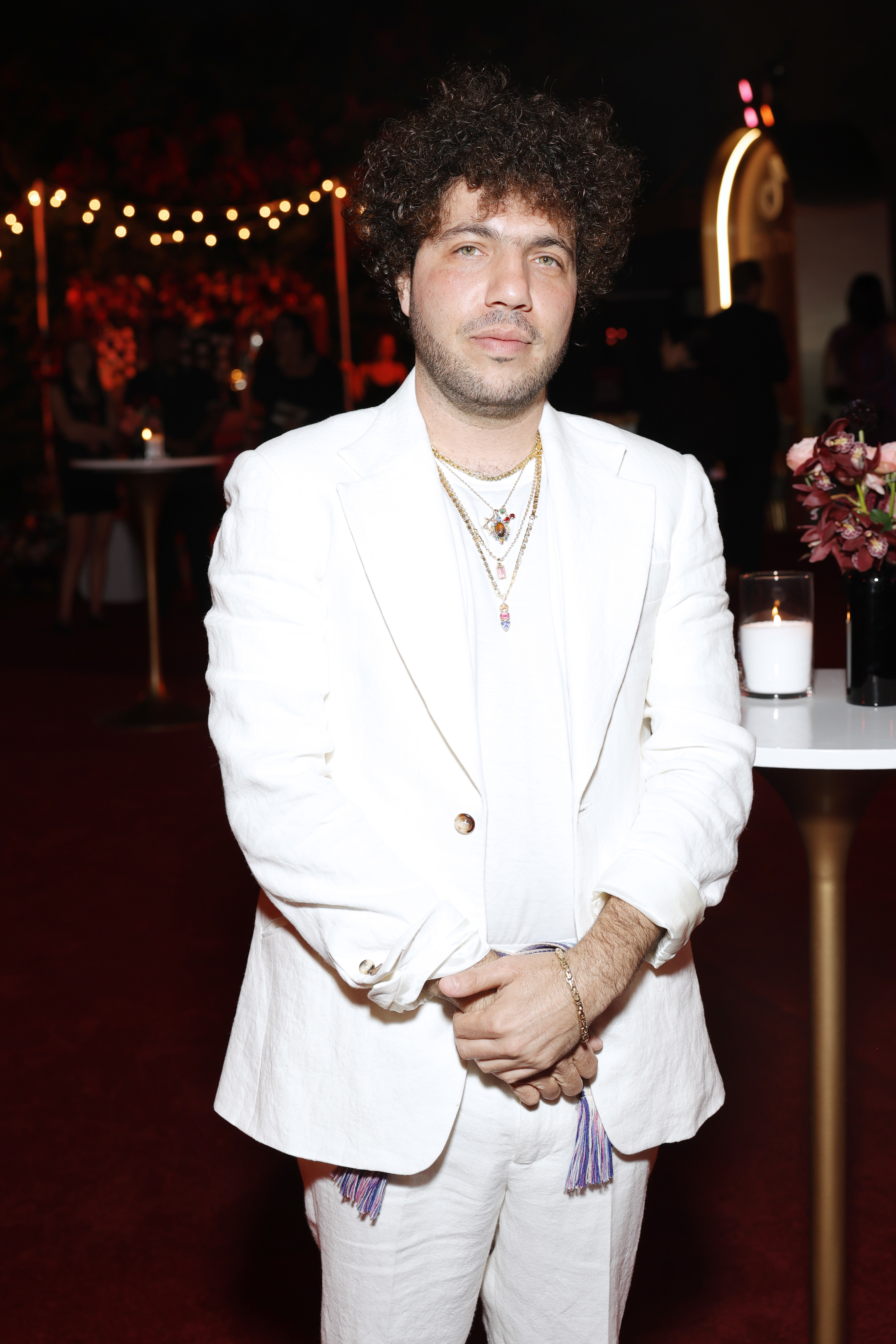 Benny Blanco stands in an event space wearing a white suit with layered necklaces. The background is adorned with string lights and guests mingling