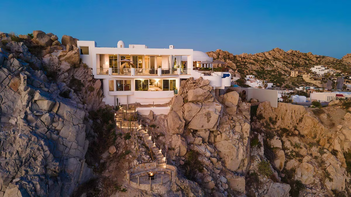 A luxurious white villa perched on a rocky cliff in a coastal area, with a lit-up winding staircase leading to the entrance. Clear sky at twilight