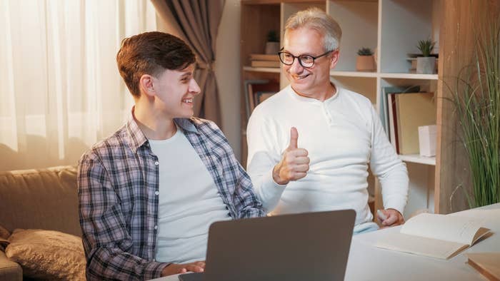 Older man wearing glasses gives a thumbs-up to a younger man while both smile near a laptop in a cozy room