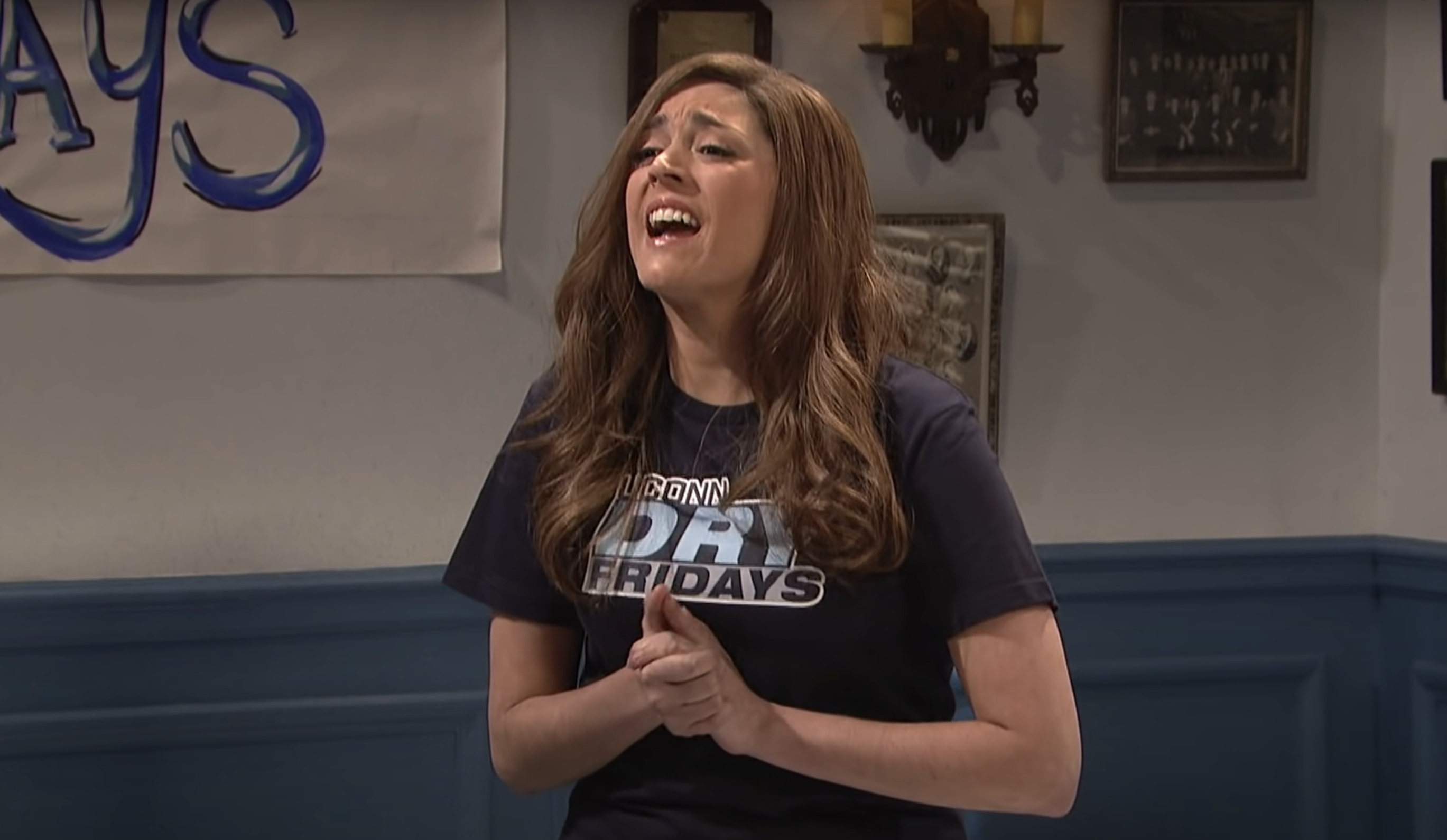 Cecily Strong as a college student, making an expressive gesture with her hands clasped, in a room with blue paneling and wall decorations