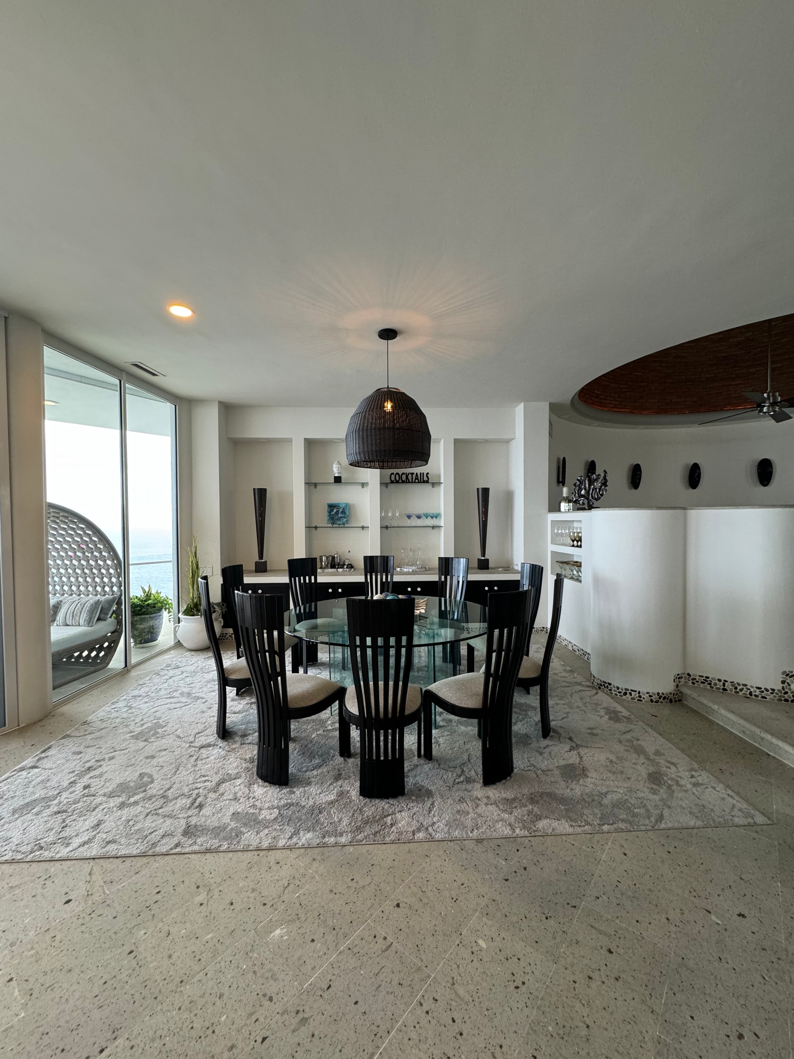 Modern, spacious dining area with glass table, black high-back chairs, and round light fixture, featuring ocean view and minimalistic decor