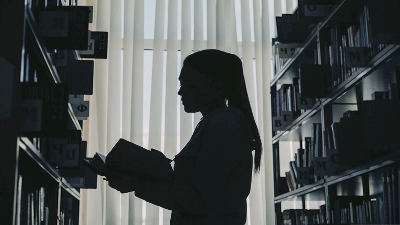 Silhouette of a person holding a book, standing between bookshelves in a library