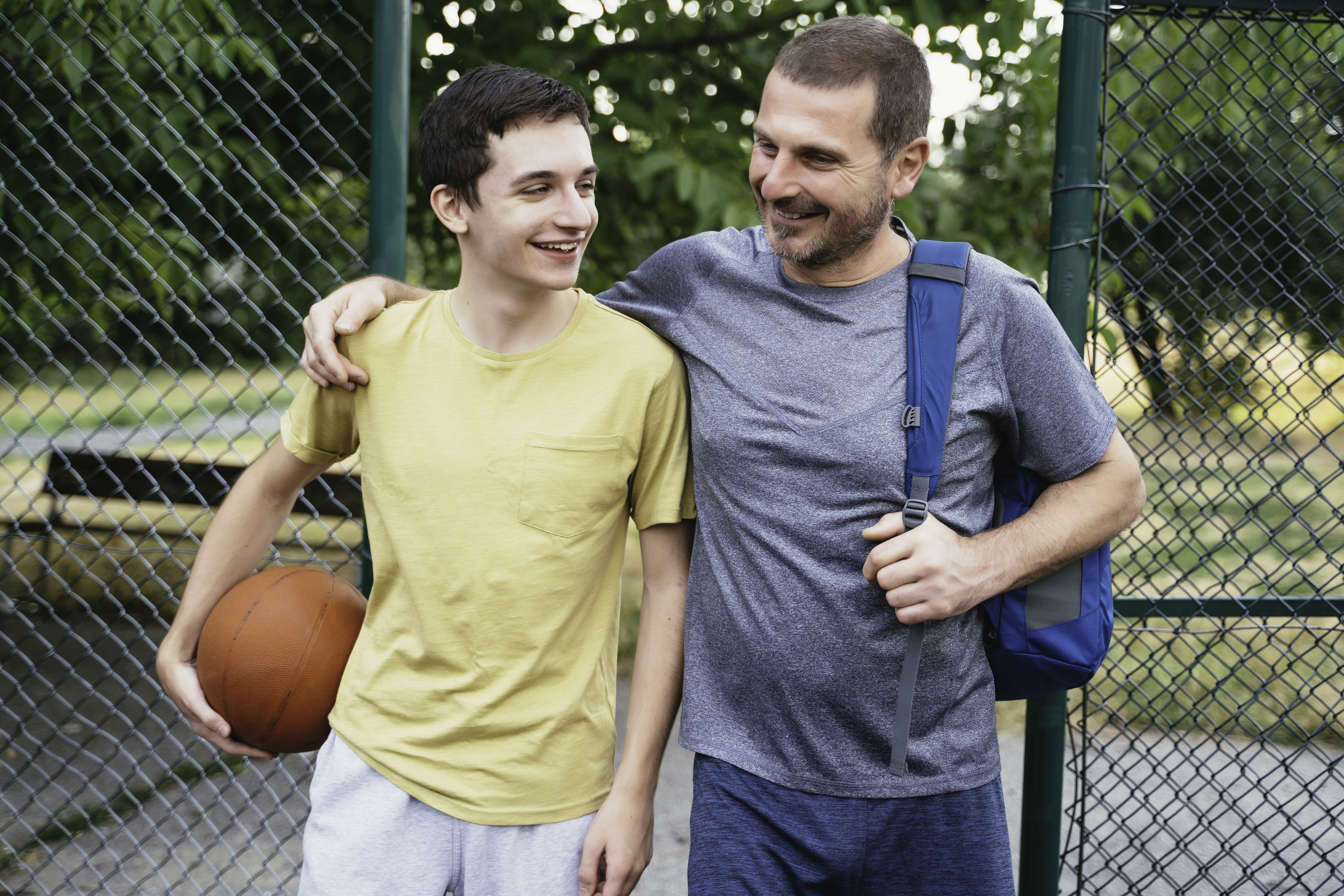 Two people smiling and walking together near a fence, one holding a basketball