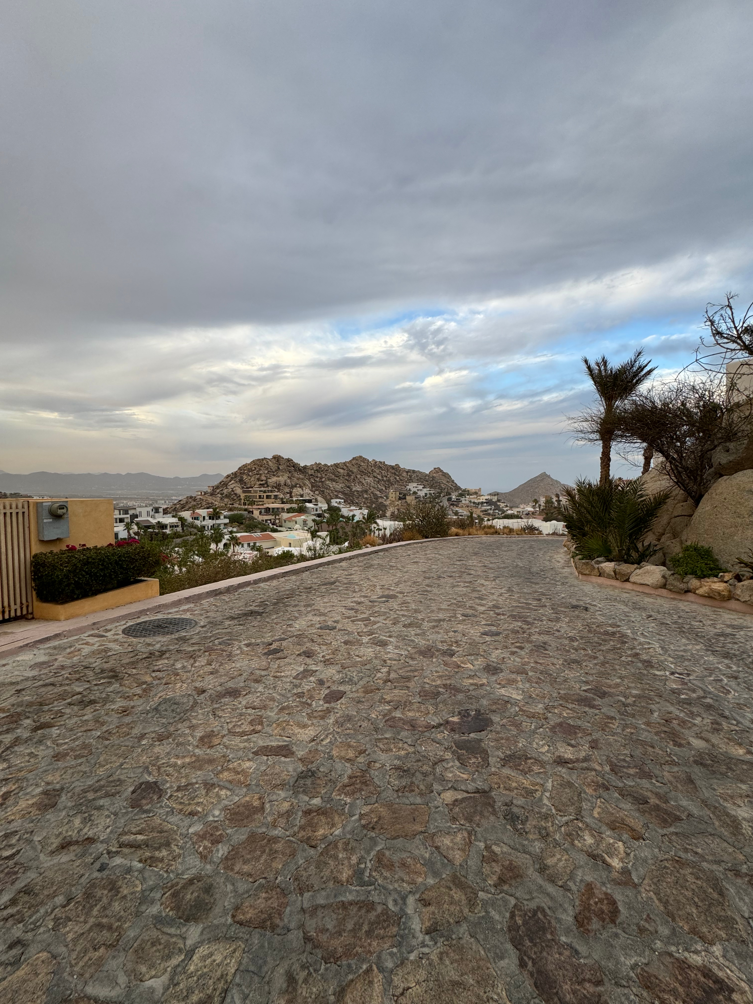 Scenic view of a cobblestone pathway in Cabo San Lucas, Mexico, with mountains and residential houses in the background under a cloudy sky